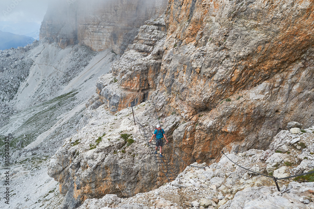 Tourist crossing a suspended wire bridge on via ferrata Cesare Piazzetta, on the way up to Piz Boe peak, on a Summer adventure tour in the Dolomites mountains, Italy. Tourist crossing a suspended wire bridge on via ferrata Cesare Piazzetta, on the way up to Piz Boe peak, on a Summer adventure tour in the Dolomites mountains, Italy.