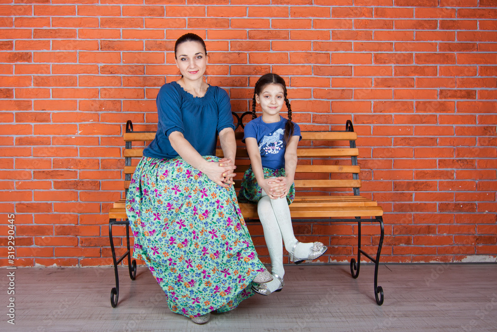 Mother and daughter and sit on the bench in front of red brick wall. Indoor photo shot