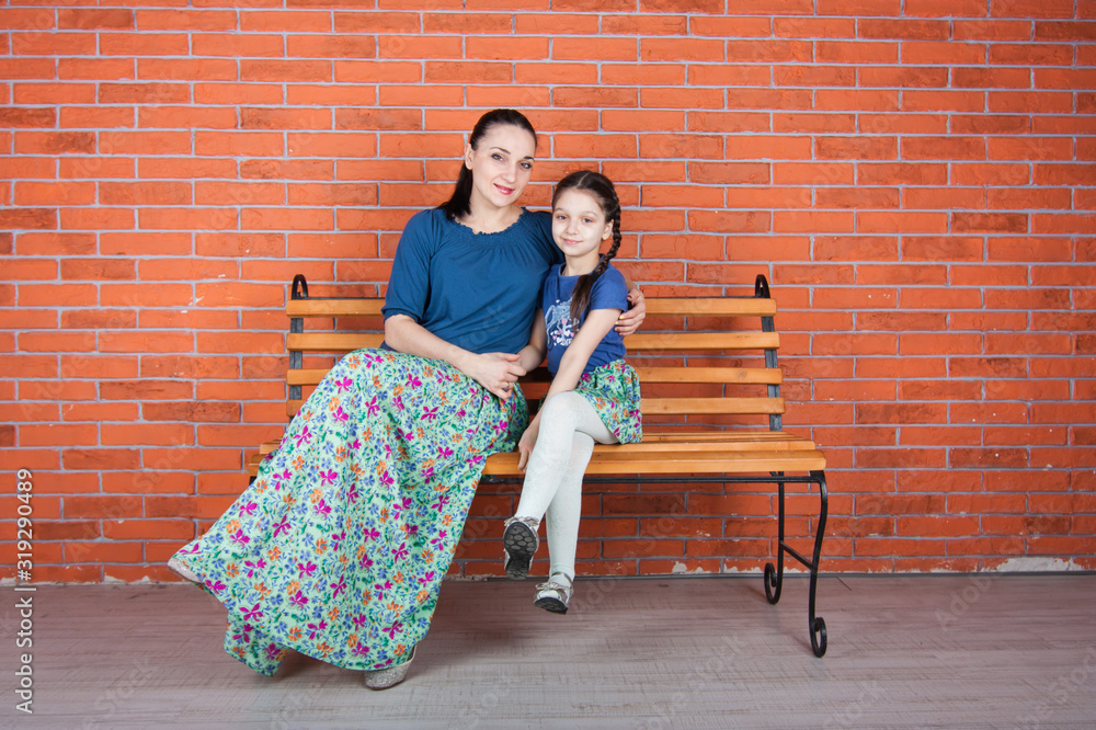 Mother and daughter in similar clothes hug and sit on the benchwith cross legs. Indoor photo shot