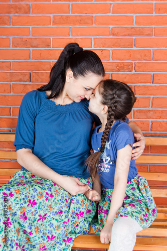 Obraz na płótnie Mother and daughter in similar clothes hug and sit on the bench in front of red brick wall. Indoor photo shot w salonie