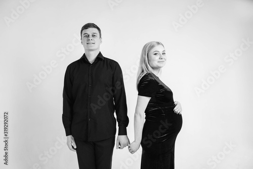 Pregnant woman and her husband holding hands in black clothes on a white background. Black and white picture. Beautiful married couple. Copy, empty space for text