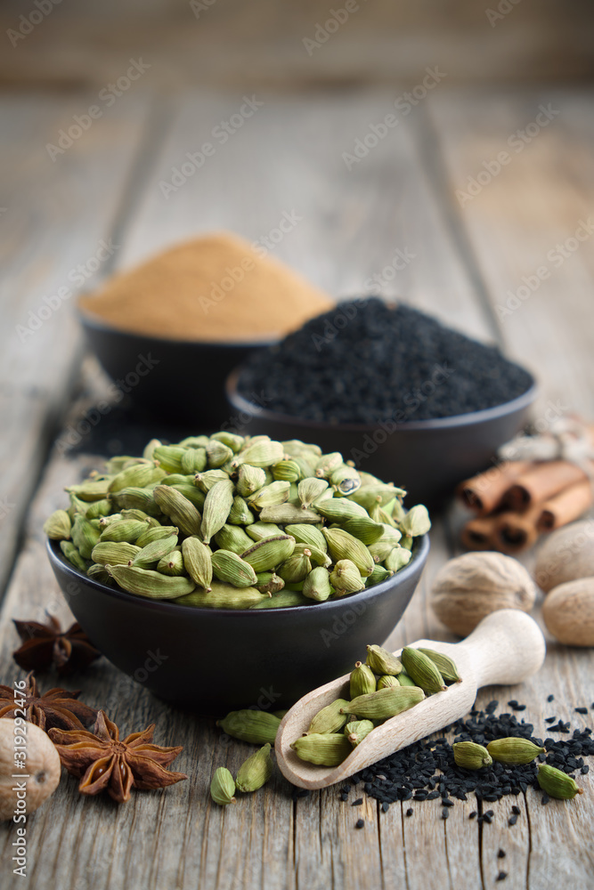 Green cardamom pods in black bowl in foreground. Aromatic spices: anise, gloves, black cumin seeds, nutmegs, cinnamon sticks, turmeric. Ingredients for cooking. Ayurveda treatments. Green cardamom pods in black bowl in foreground. Aromatic spices: anise, gloves, black cumin seeds, nutmegs, cinnamon sticks, turmeric. Ingredients for cooking. Ayurveda treatments.