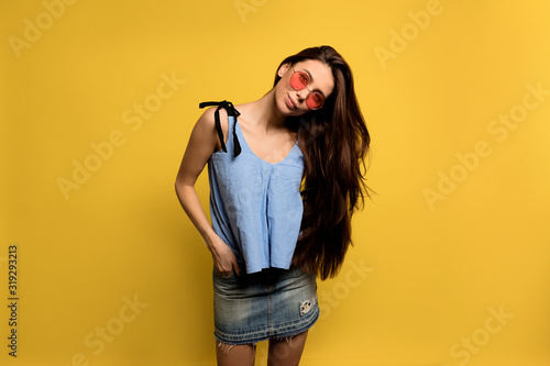 Stylish hipster girl with long brown hair wearing blue t-shirt and denim short skirt in round pink glasses posing over yellow background