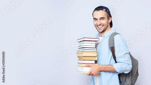Happy student guy with backpack carries a stack of books on gray background, copy space, 16:9