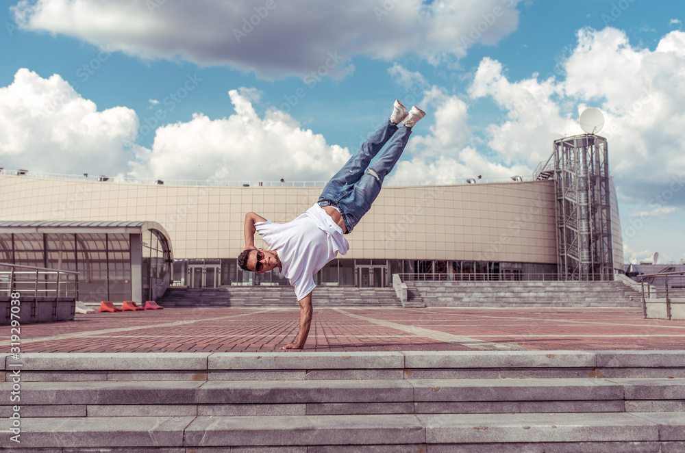 Fototapeta premium man standing on one arm, young guy dancer, summer city, dancing street dances, break of movement, modern youth style. Fashion fitness sport. Free space for copy text. Step cloud background.