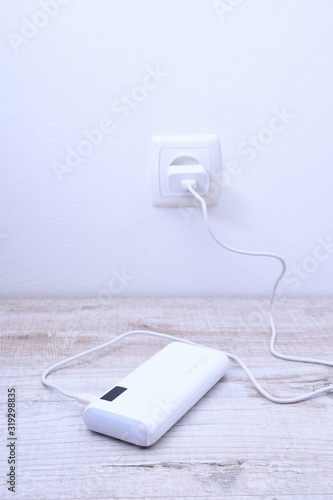 White phone power bank plugged in the socket on the wall for charging on wooden background