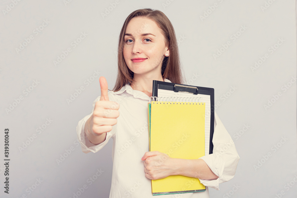 Happy woman student with notebooks shows thumb up. Concept of education. Portrait, grey background, copy space, slogan, toned Happy woman student with notebooks shows thumb up. Concept of education. Portrait, grey background, copy space, slogan, toned