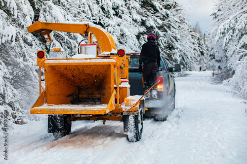 A wide view of an arboriculturist, tree surgeon or landscape gardener, towing a mobile wood chipper with a pickup truck down a snow covered street photo