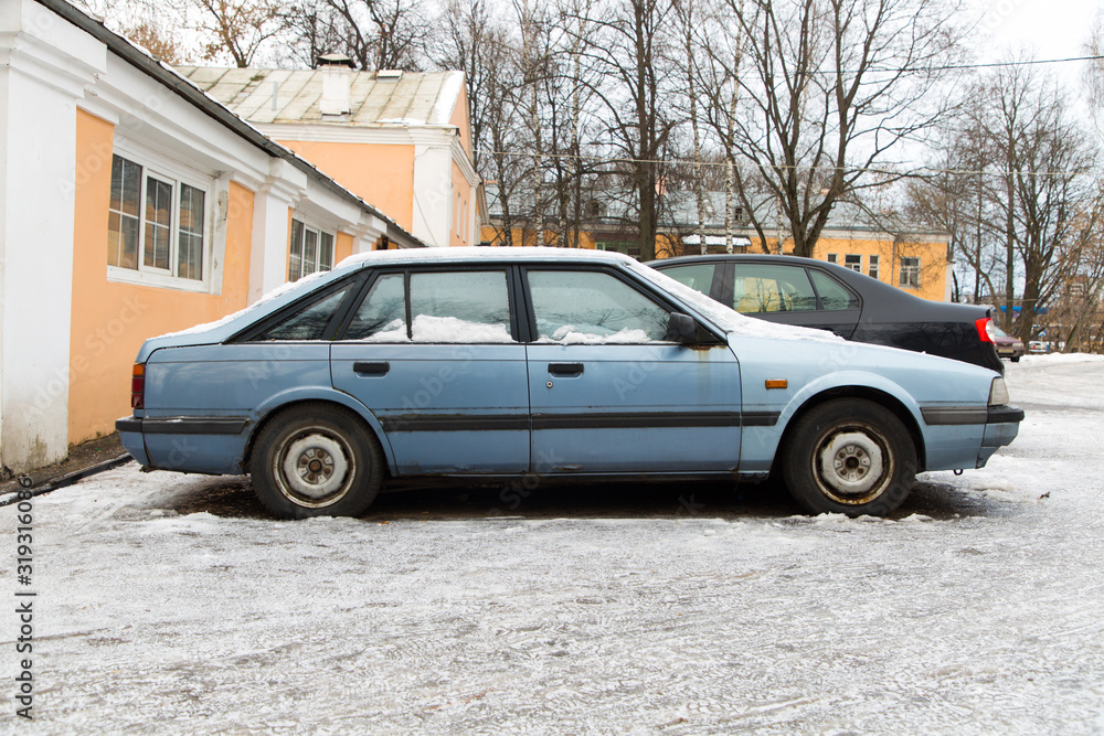 An old car of blue color stands in a parking lot under the snow, against the background of old houses