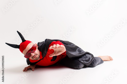 Fotografia Conceptual portrait of defeated and helpless devil man in black red vampire costume asking for help and mercy while lying on ground in studio and reaching his arm out