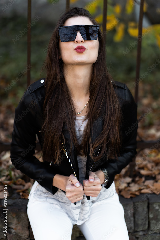 Outdoor fashion portrait of stylish young woman having fun, emotional face , laughing, Urban city street style.Girl wearing white pants,t-shirt, black leather jacket , sunglasses and dark hat Fashion.