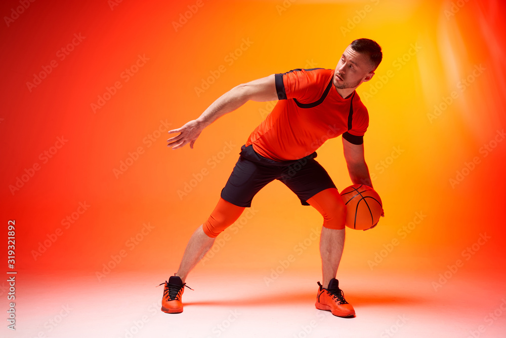 Young athletic basketball player in attack position holding ball with one hand on orange and red background