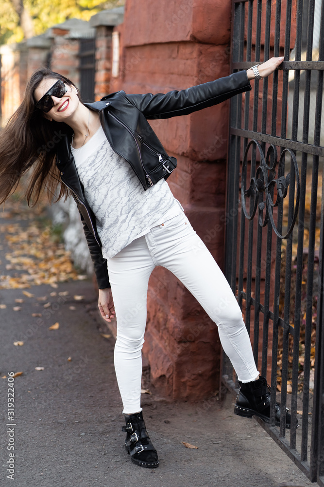 Outdoor fashion portrait of stylish young woman having fun, emotional face , laughing, Urban city street style.Girl wearing white pants,t-shirt, black leather jacket , sunglasses and dark hat Fashion.