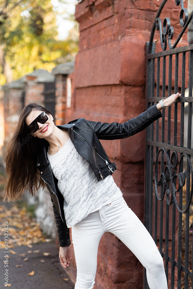 Outdoor fashion portrait of stylish young woman having fun, emotional face , laughing, Urban city street style.Girl wearing white pants,t-shirt, black leather jacket , sunglasses and dark hat Fashion.