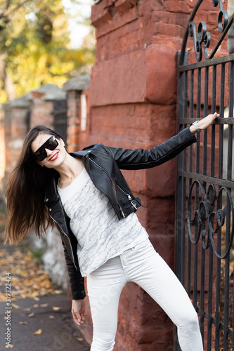 Obraz na płótnie Outdoor fashion portrait of stylish young woman having fun, emotional face , laughing, Urban city street style.Girl wearing white pants,t-shirt, black leather jacket , sunglasses and dark hat Fashion. w salonie