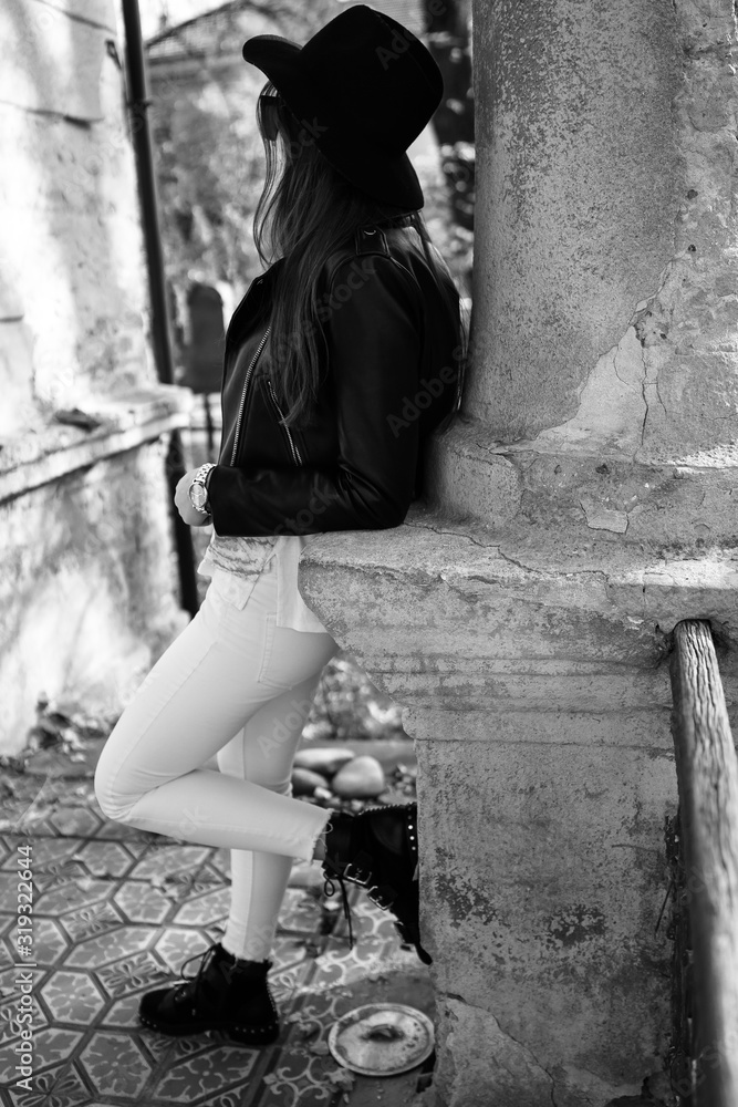 Outdoor photo of brunette lady posing on tree background in autumn day.Fashion street style portrait. Girl wearing white pants,t-shirt, black leather jacket , sunglasses and dark hat .Fashion concept.