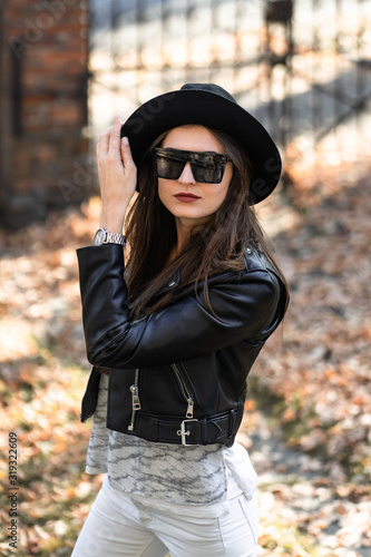 Outdoor photo of brunette lady posing on tree background in autumn day.Fashion street style portrait. Girl wearing white pants,t-shirt, black leather jacket , sunglasses and dark hat .Fashion concept.