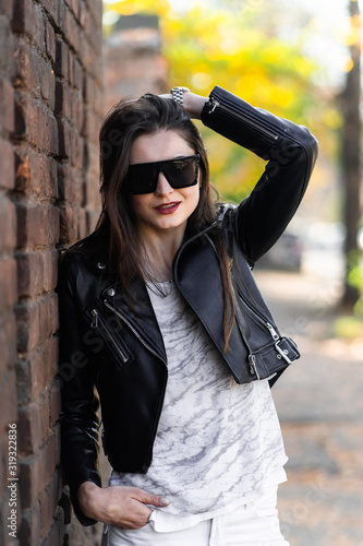 Obraz na płótnie Outdoor photo of brunette lady posing near brick wall in autumn day.Fashion street style portrait. Girl wearing white pants,t-shirt, black leather jacket , sunglasses and dark hat .Fashion concept. w salonie