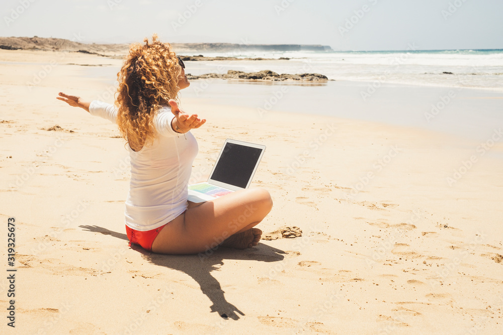 Happy and free beautiful woman sit down at the beach with laptop computer on the legs - concept of digital nomad and modern technology - connected everywhere people