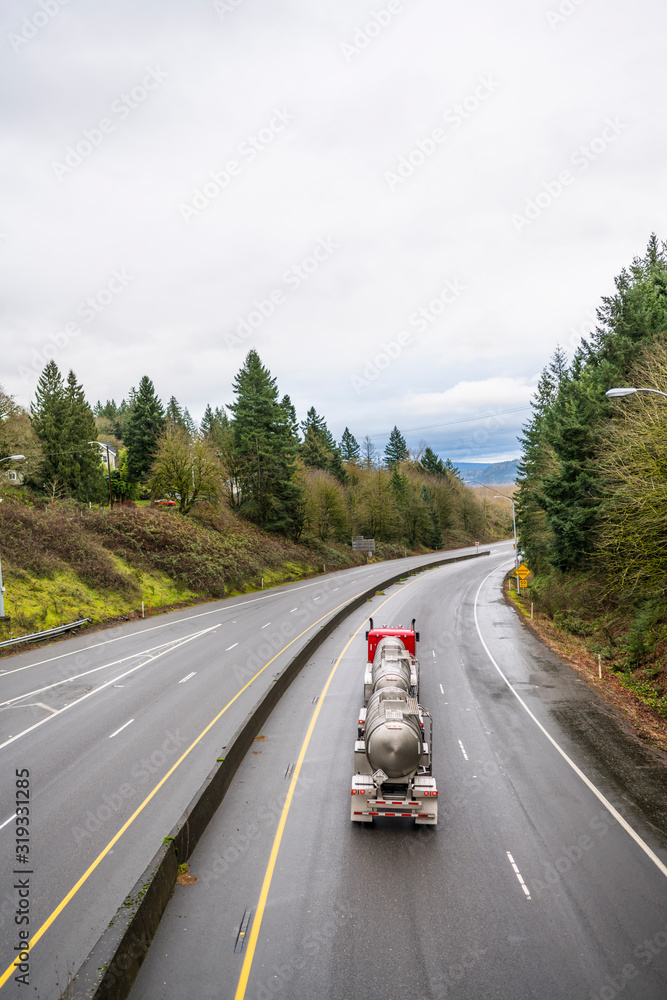 Red big rig semi truck transporting Chemical and flammable liquids in tank semi trailer running on the divided wet slippery road