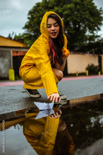young woman in yellow raincoat playing with a paper boat and a red balloon in a puddle, girl having fun on the street near the gutter