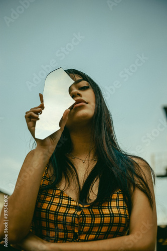 Girl in orange print dress holding piece of broken mirror reflecting sky clouds in front of her face photo