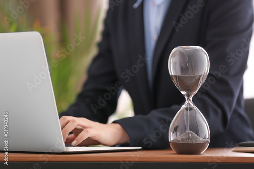 Hourglass near woman working on laptop at table in office. Time management concept