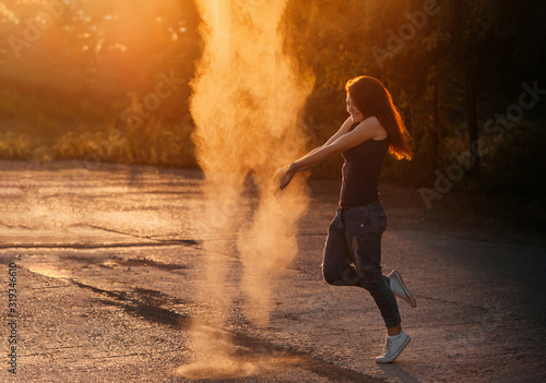 a slender girl with long red hair throws up magic dust or sand, whirls and laughs in the sunset backlight, a pillar of road dust near a person