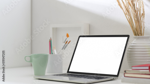 Cropped shot of simple artist workspace with blank screen laptop, coffee cup, painting brush and decorations on white table © bongkarn