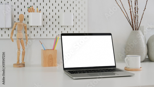 Cropped shot of modern designer workspace with mock up laptop, wooden figure, pencils and ceramic vase on white table © bongkarn