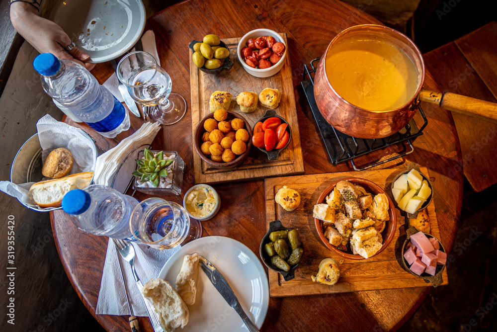 Delicious cheese fondue with dips, pickles, olives, pork, croutons, sausage and cherry tomatoes seen from above.