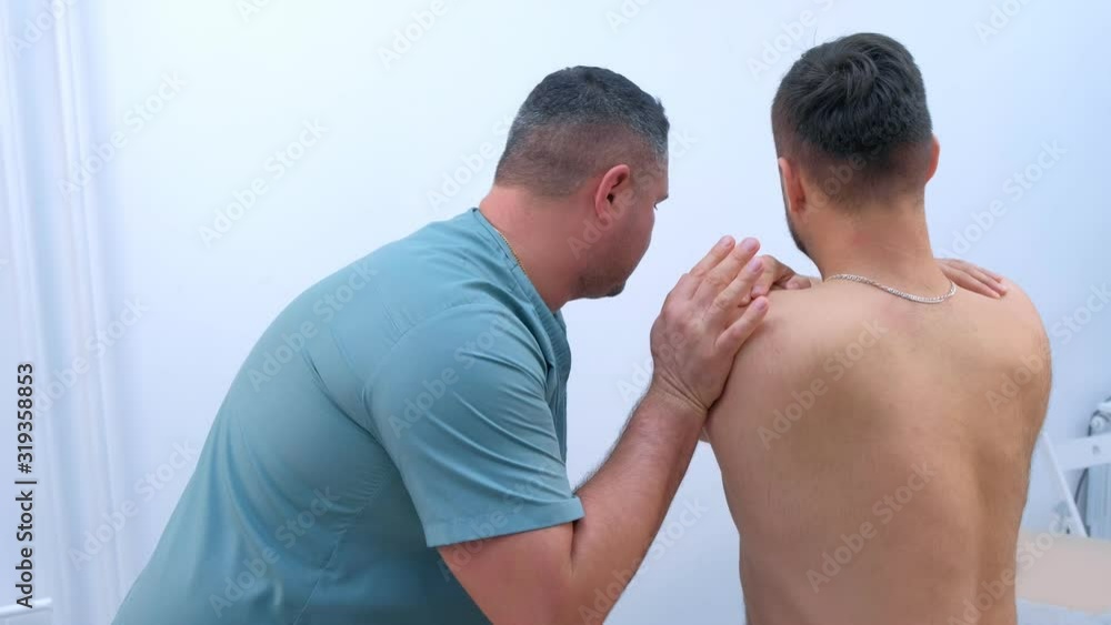 Doctor chiropractor osteopath treats patient's shoulder in clinic on rehabilitation therapy. Man with neck torso sitting back to camera. Manual therapist physician appointment and healing treatment. Doctor chiropractor osteopath treats patient's shoulder in clinic on rehabilitation therapy. Man with neck torso sitting back to camera. Manual therapist physician appointment and healing treatment.