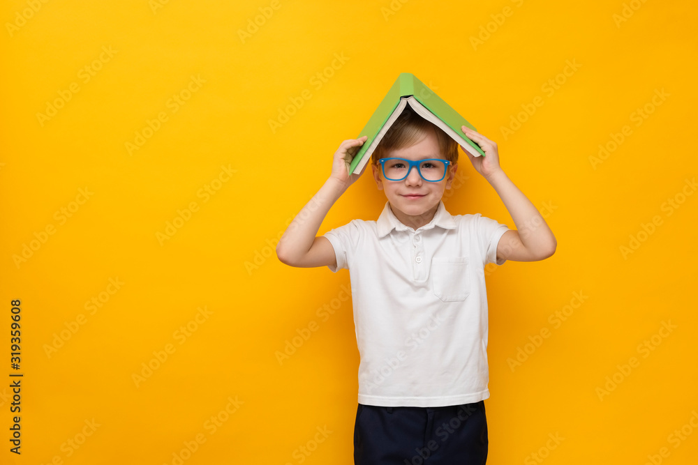 Cute little school boy in glasses holding book on his head on yellow background, , copy space Cute little school boy in glasses holding book on his head on yellow background, , copy space