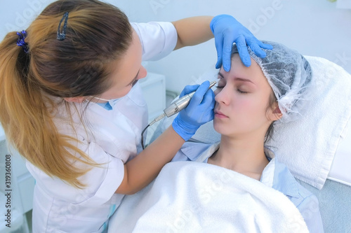 Cosmetologist making eyebrows microblading procedure in beauty salon for girl using tattoo apparatus. Beautician in gloves is doing permanent eyebrow makeup for woman. Beauty industry concept.