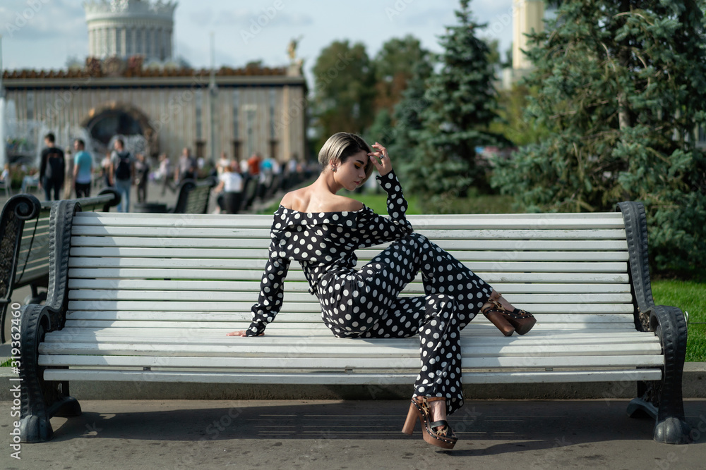 posing on a park bench. beautiful European model in a black trouser suit with white spots, pants and a shirt, high heel. Short haircut, bright makeup, long legs. female lessons posing