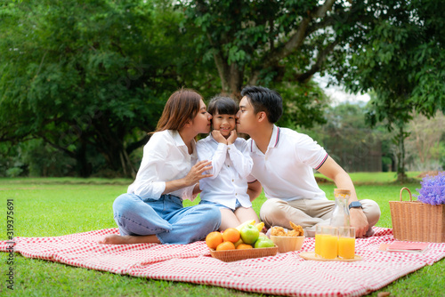 Asian teen family happy holiday picnic moment in the park with father, mother kissing daughter looking at camera and smile to happy spend vacation time togerter in green garden with friut and food..