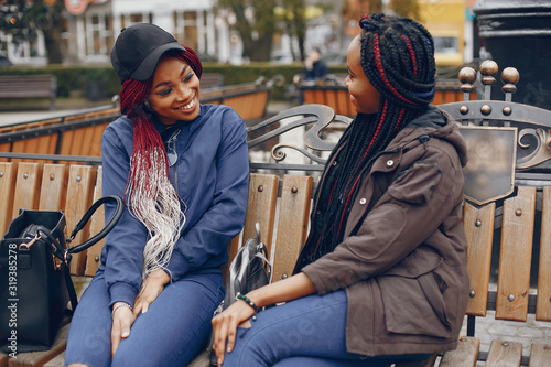 two beautiful and stylish dark-skinned girls with long hair sitting on the bench in a autumn city