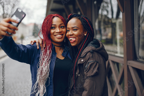 two beautiful and stylish dark-skinned girls with long hair stnding near building in a autumn city and make a selfie