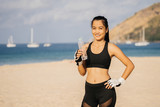 Portrait of a sporty asian girl with a bottle of clear water in sportswear on a tropical beach. Sports lifestyle and health.