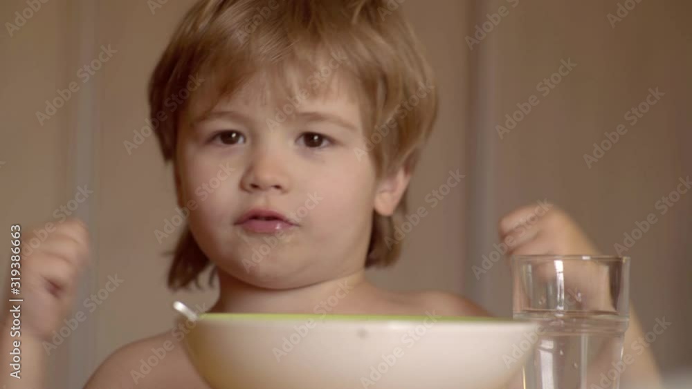Little boy sitting at the table and eating milk snack. Baby eats food itself with spoon. Portrait of sweet little laughing baby boy with blonde hair eating from plate holding spoon close-up. Little boy sitting at the table and eating milk snack. Baby eats food itself with spoon. Portrait of sweet little laughing baby boy with blonde hair eating from plate holding spoon close-up.