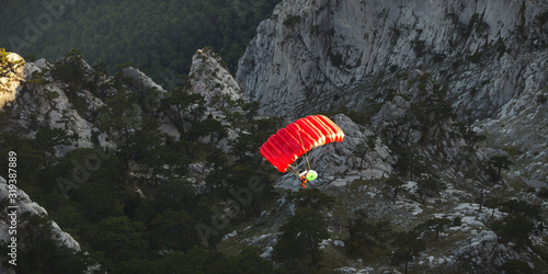 Base jumper under a bright red parachute flies against a background of a dark rocky landscape after jumping from the top of a mountain, view from above. Panorama.