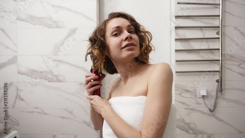 Smiling young woman in big white bath towel styling her hair with a curling iron in front of a mirror. A girl after shower makes a hairstyle in the bathroom. Front view. Marble wall on background photo