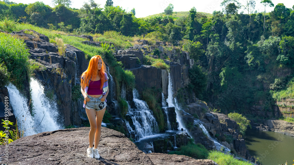 Beautiful young red haird girl staying near amazing  waterfall in Da Lat city Vietnam.Traveler girl with long red hair looking to beautiful Pongour  waterfall.