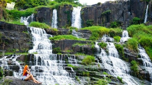 Beautiful young red haird girl sitting near amazing waterfall in Da Lat city Vietnam.Traveler girl with long red hair looking to beautiful Pongour waterfall.
