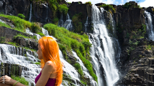 Beautiful young red haird girl sitting near amazing  waterfall in Da Lat city Vietnam.Traveler girl with long red hair looking to beautiful Pongour  waterfall. photo