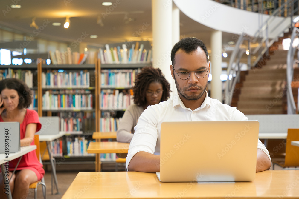 Front view of focused man typing on laptop. Handsome young student sitting at library and using modern device. Technology concept Front view of focused man typing on laptop. Handsome young student sitting at library and using modern device. Technology concept