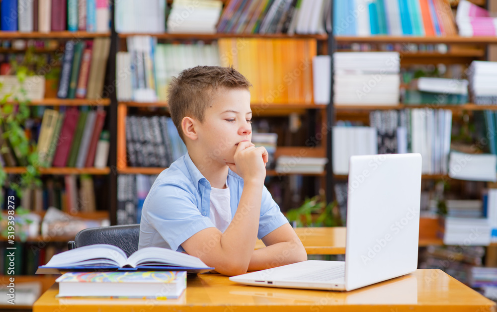 A boy sits in a library at a table, works on a laptop, looks thoughtfully at the screen