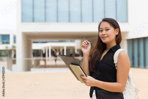 Female student holding folder and smiling at camera. Beautiful cheerful young Asian woman holding pen, folder and book on street. Education concept