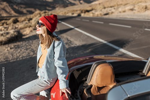 Woman standing on the roadside near the convertible car, traveling on the dessert valley