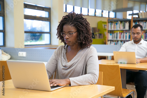 Concentrated young woman working with laptop at library. Focused African American woman typing on laptop. Technology concept